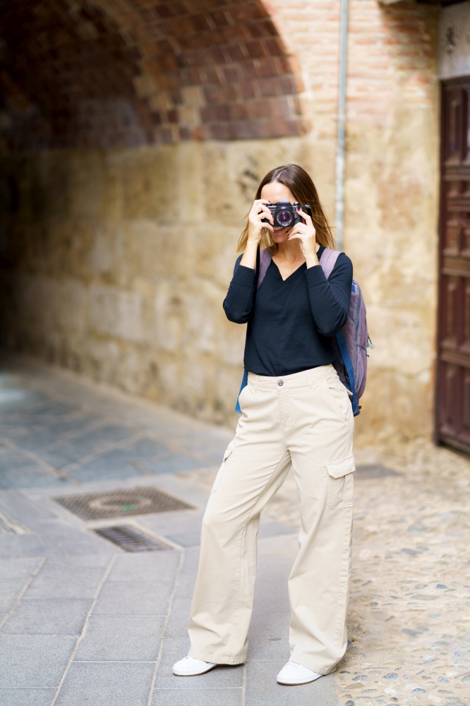 Full body of young female tourist in casual outfit with backpac,standing near aged brick building with arched passage and taking pictures on photo camera during sightseeing trip. Content woman taking photos on camera near arched passage in old town