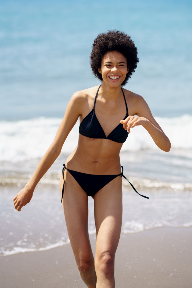 Delighted African American female in swimwear strolling on wet sandy beach near waving sea during summer vacation in tropical resort. Joyful black woman in bikini walking along seaside