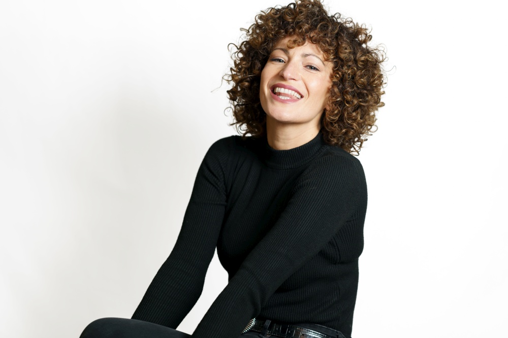 Positive young female with curly hair smiling with opened mouth and looking at camera while sitting with hands near leg against white background. Smiling woman with curly hair sitting against white wall