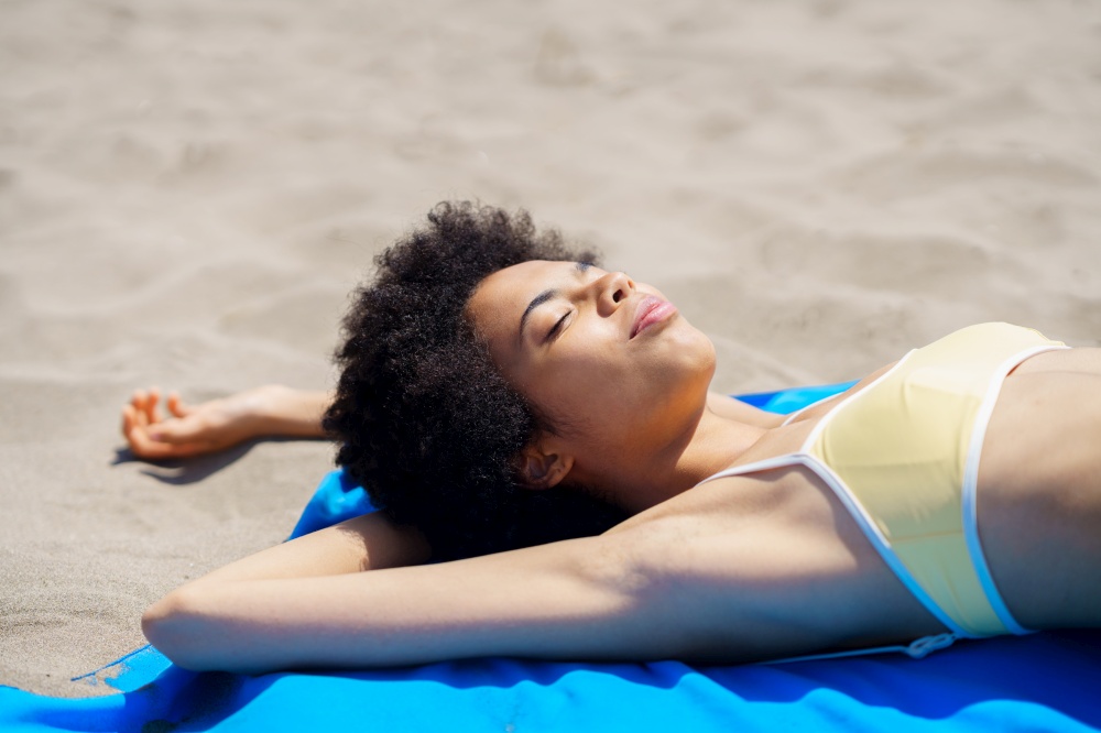 Happy young African American female tourist with curly hair in trendy bikini lying on sandy beach with closed eyes while sunbathing during summer holidays. Relaxed ethnic lady sunbathing on sandy shore on summer day