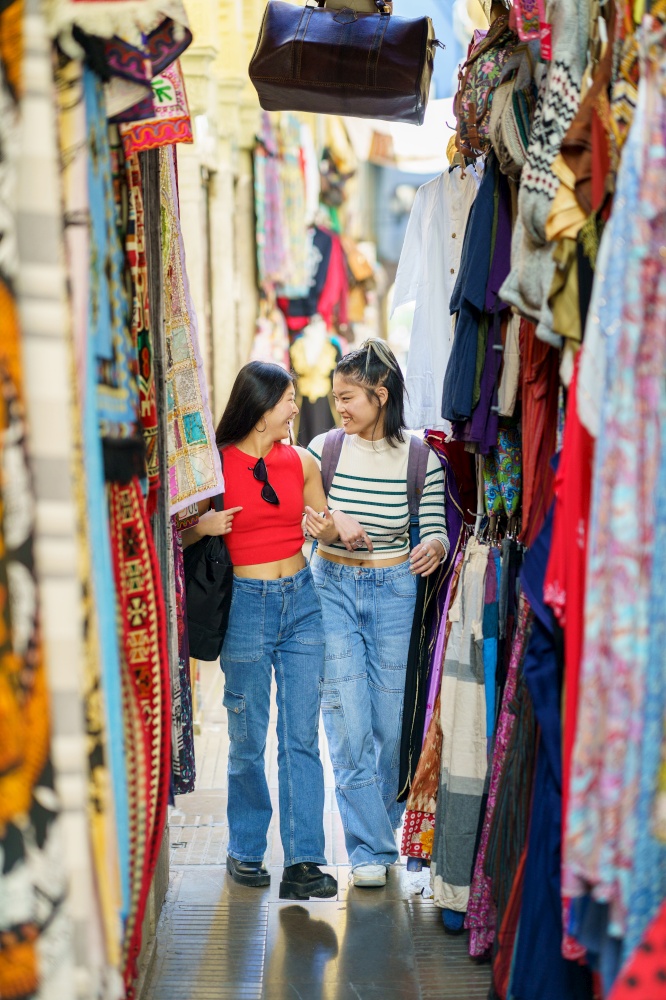 Smiling young women friends in casual clothes walking along colorful clothing store on street market in city during summer vacation. Two Chinese women tourists shopping clothes in street bazaar