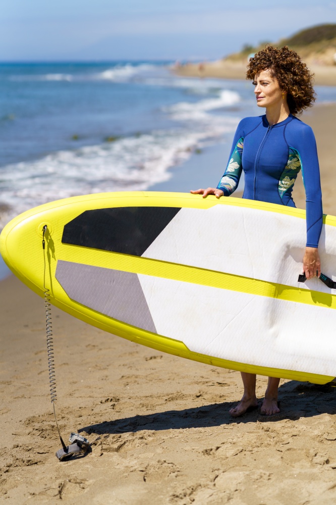 Side view of smiling young female redhead in swimsuit standing on sandy beach with paddleboard in hands and waiting while looking away against blurred seacoast. Young woman standing with SUP board on sandy beach
