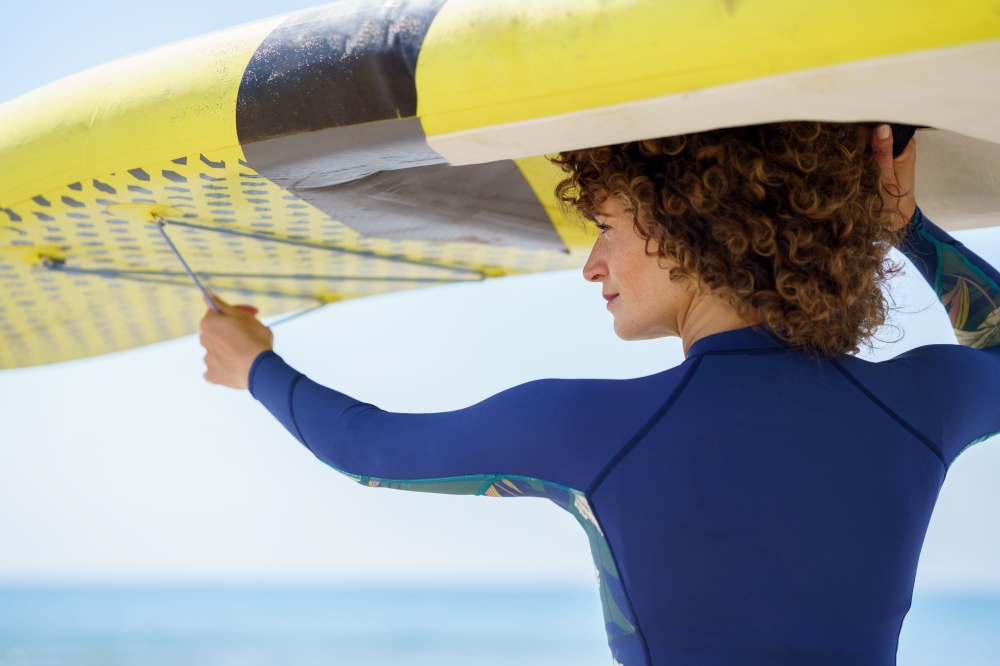 Back view of positive curly haired female in trendy swimwear with colorful paddleboard on background of sea looking away in bright daylight. Happy woman in curly hair lifting yellow SUP board