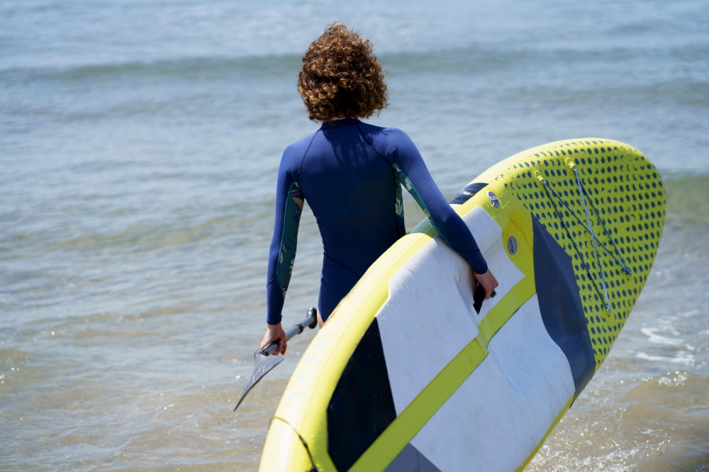 Back view of unrecognizable female surfer in wetsuit with SUP board and paddle standing in water at beach in rippling blue sea at daylight. Unrecognizable surfer with paddleboard in sea