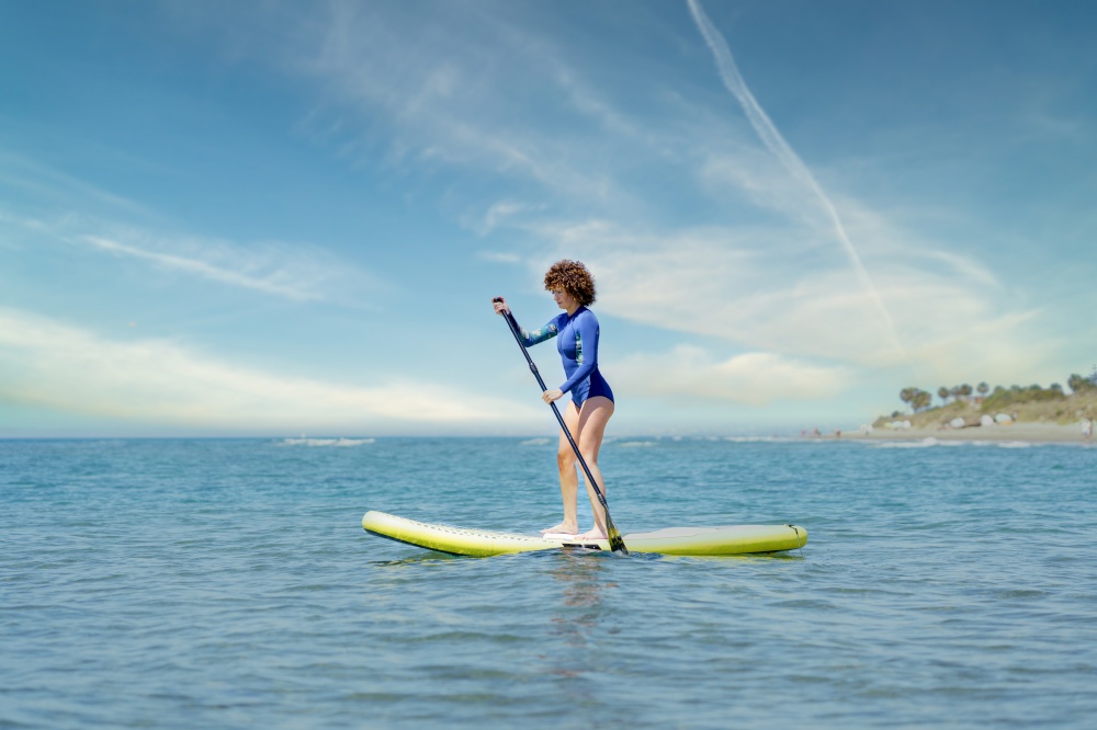 Focused female surfer in blue wetsuit standing on SUP board and paddling in sea against cloudy blue sky in sunny summer day. Woman on paddleboard in sea