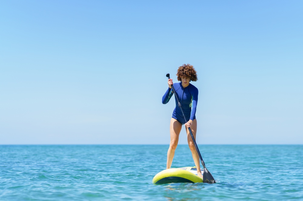 Careful female surfer in wetsuit standing on SUP board and paddling blue sea against cloudless blue sky in sunny summer day. Curly hair woman with paddleboard in sea