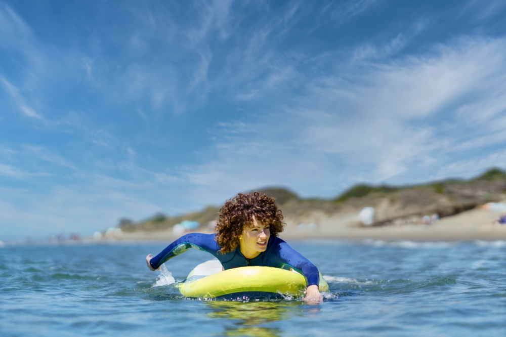 Young curly haired woman in wetsuit lying on standup SUP board paddling with hands in sea against blue sky on sunny summer day. Fit woman on paddleboard rowing with hands in ocean