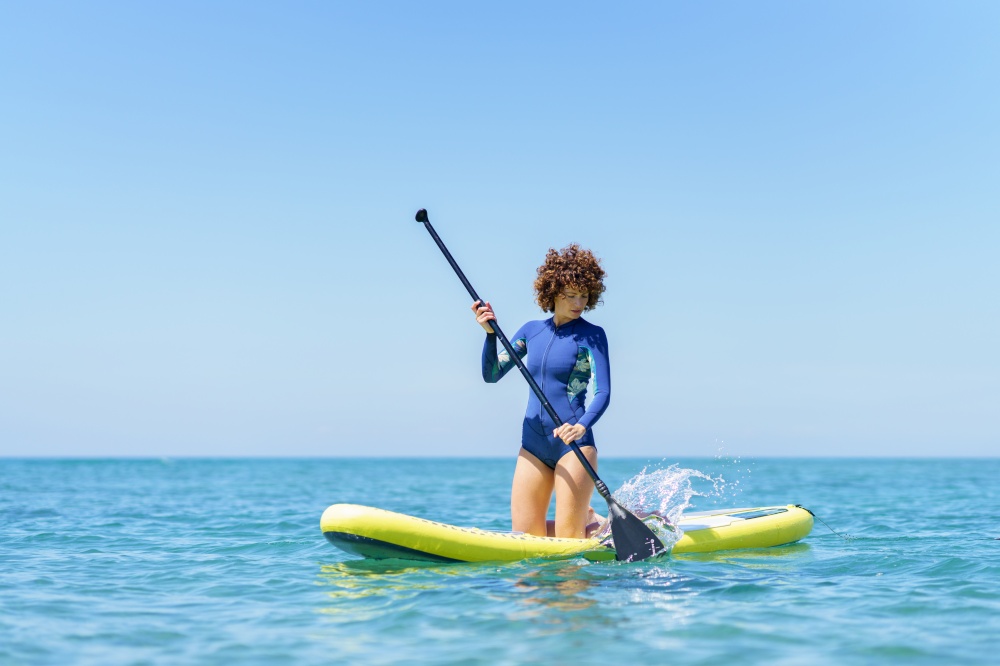 Focused curly hair woman in wetsuit with paddle kneeling on SUP board while paddling on blue sea under blue cloudless sky. Woman with paddle on paddleboard in sea