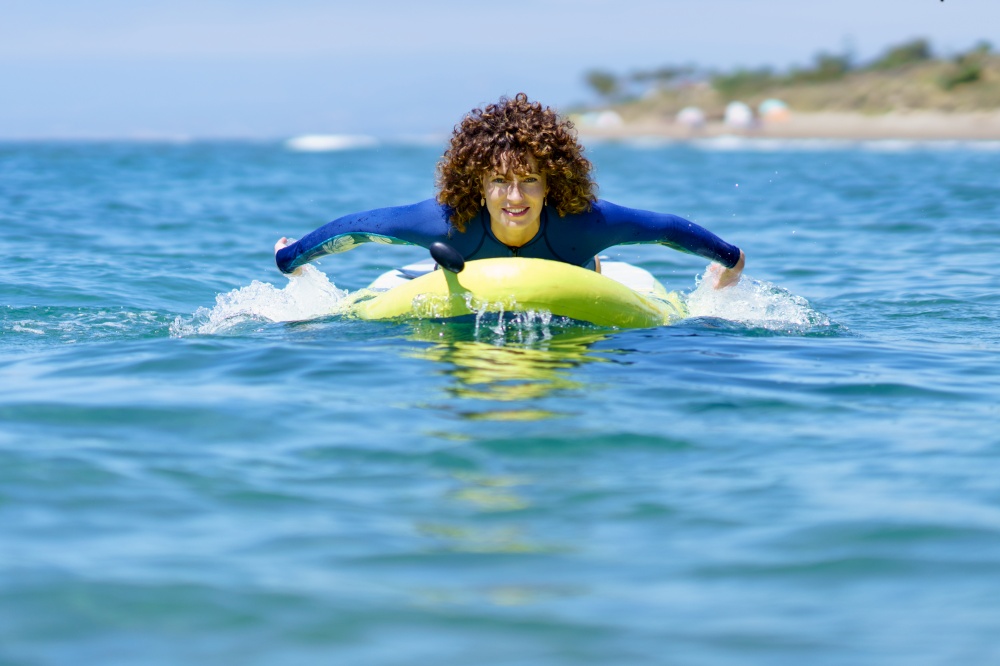Content curly haired female in wetsuit pulling SUP board in rippling seawater and looking at camera in daylight. Woman swimming on paddleboard in sea