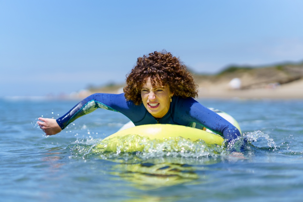 Young curly haired female surfer in wetsuit lying on SUP board and paddling with hands floating over sea against blue sky. Determined woman on paddleboard rowing with hands in ocean