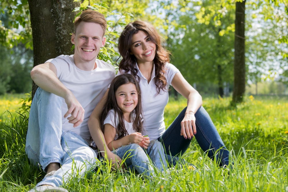 Portrait of happy family sitting on grass under a tree in park on a sunny day. Portrait of happy family in park