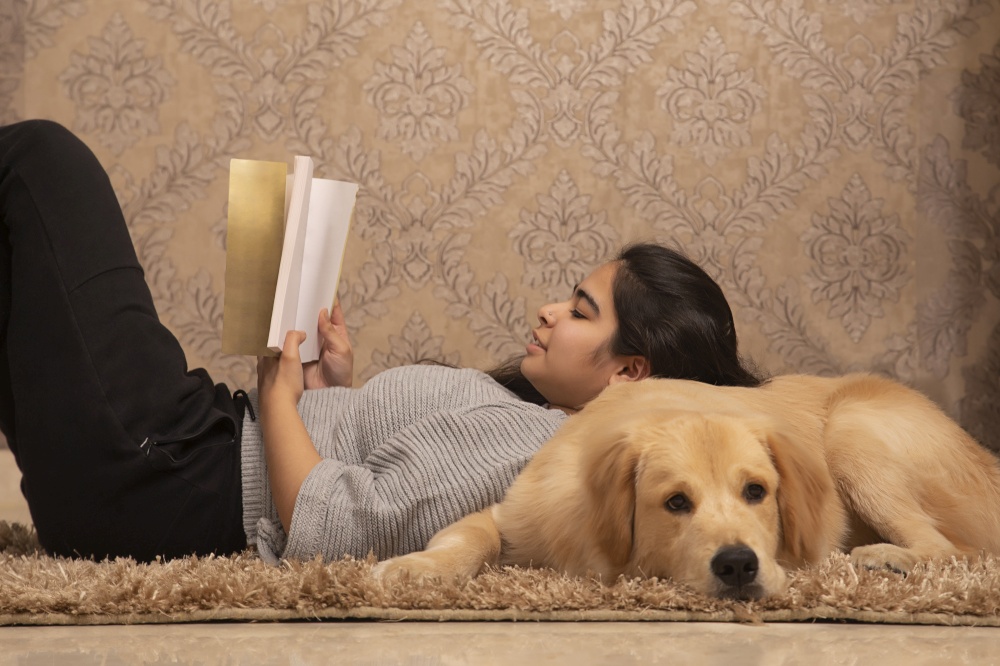 A YOUNG GIRL RESTING ON FLOOR AND READING BOOK WITH PET DOG BESIDES