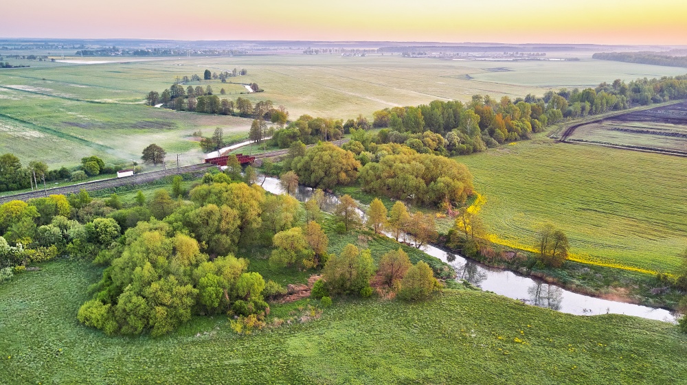 Spring morning landscape. Railroad bridge crossing calm river in blooming meadows. Aerial rural view from above. Agricultural yellow green colza fields. Dandelion blossom scene. Europe, Belarus