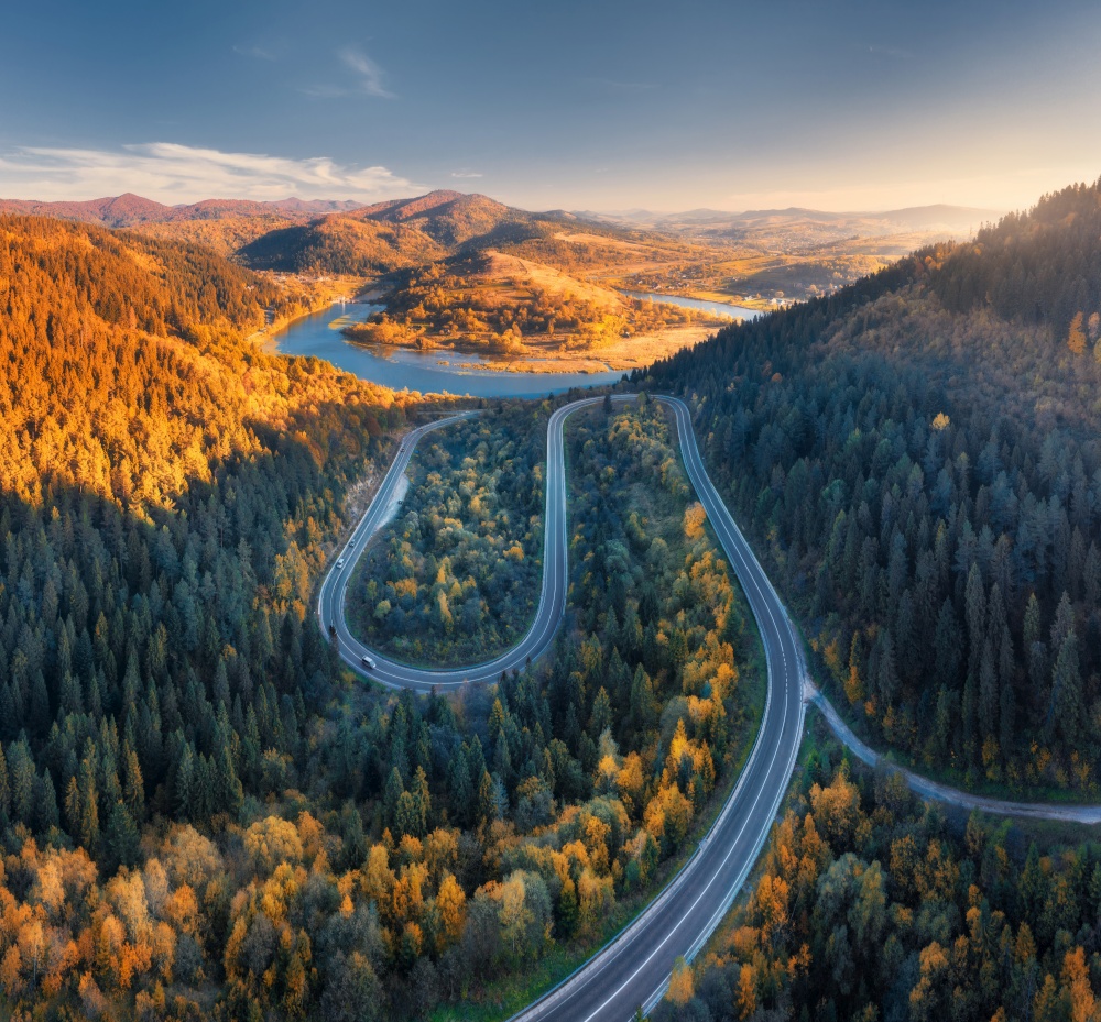 Aerial view of mountain road in colorful forest at sunset in autumn in Ukraine. Top view from drone of road in woods in fall. Beautiful landscape with highway in hills, river, pine trees, mountains. Aerial view of mountain road in colorful forest at sunset in autumn