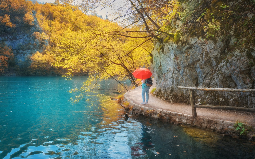 Woman with red umbrella on path under yellow trees near beautiful lake in rain in autumn. Plitvice Lakes, Croatia. Colorful landscape with girl, trail, orange forest, azure water in park in fall