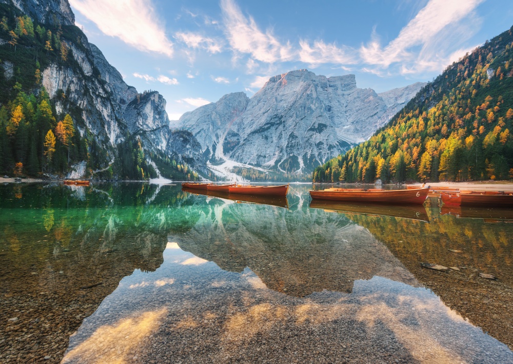 Beautiful wooden boats on Braies lake at sunrise in autumn in Dolomites, Italy. Landscape with rocky mountains, water, reflection, trees with orange leaves in fall at dawn. Travel in italian alps