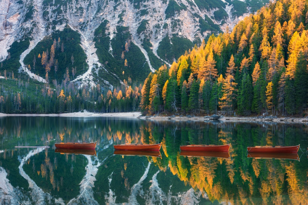 Red wooden boats on Braies lake at sunrise in autumn in Dolomites, Italy. Landscape with fall forest, mountains, water with reflection, trees with colorful foliage. Travel in italian alps. Nature