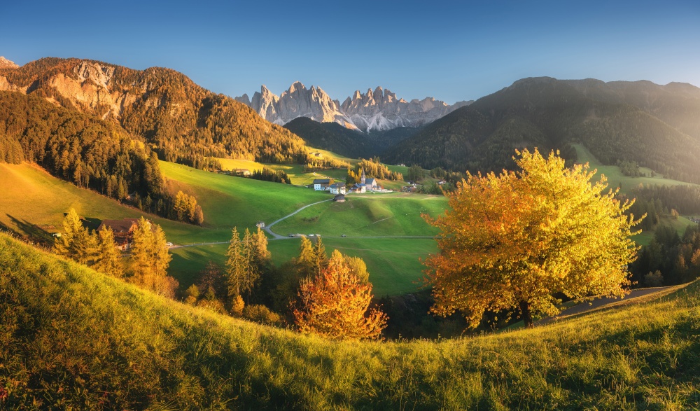 Landscape with colorful trees, village with houses, church, green meadows, rocks, blue sky at sunset in autumn in Dolomites, Italy. Santa Maddalena in mountain valley. St. Magdalena in fall. Landscape
