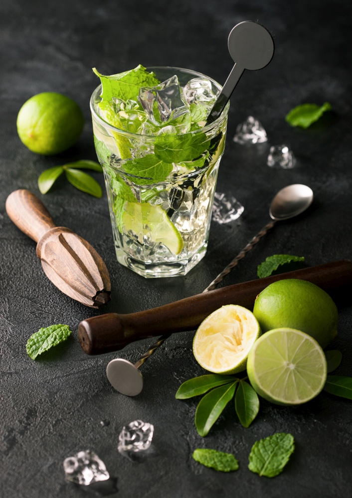 Glass of Mojito cocktail with ice cubes mint and lime on black board with spoon and muddler and fresh limes and cane sugar with wooden squeezer. Best summer cocktail