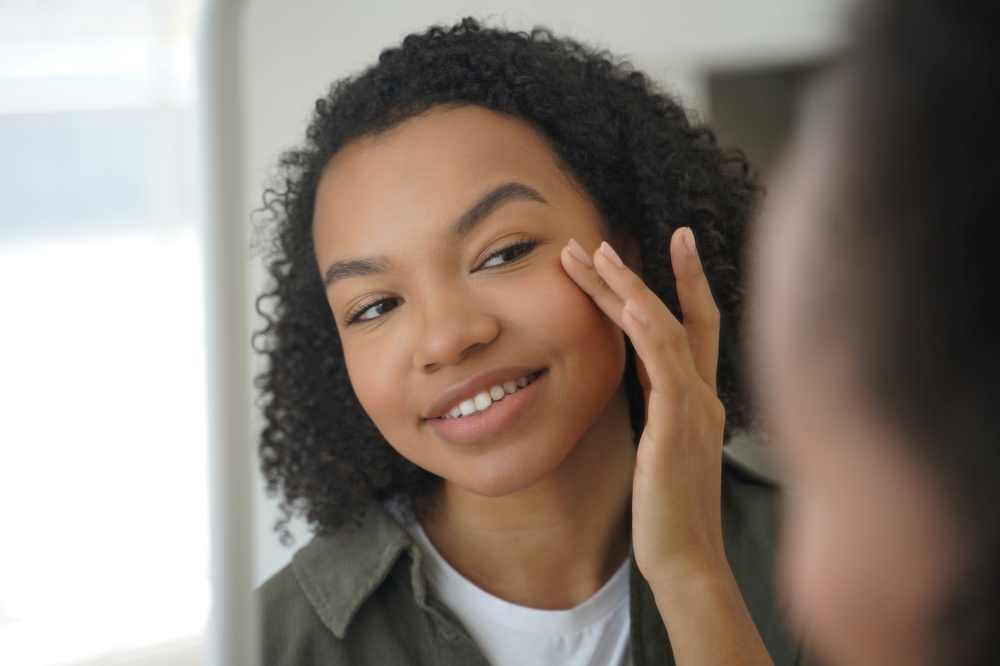 Biracial young girl looks at mirror, touches delicate under eye skin, feels happy after moisturizing mask. Mixed race woman apply daily facial cream. Natural beauty and skin care concept.. Biracial young girl looks at mirror, applying moisturizing facial cream. Natural beauty, skincare
