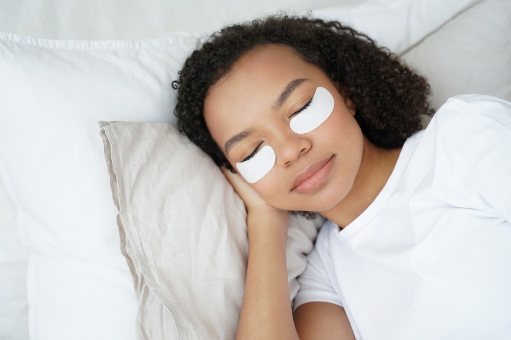 Young biracial girl with under eye patches on face relaxing lying in bed with closed eyes. Pretty woman using undereye skin beauty masks, preventing bags, reducing wrinkles. Morning skincare routine.. Young biracial girl with under eye patches on face relax lying in bed. Morning skincare routine