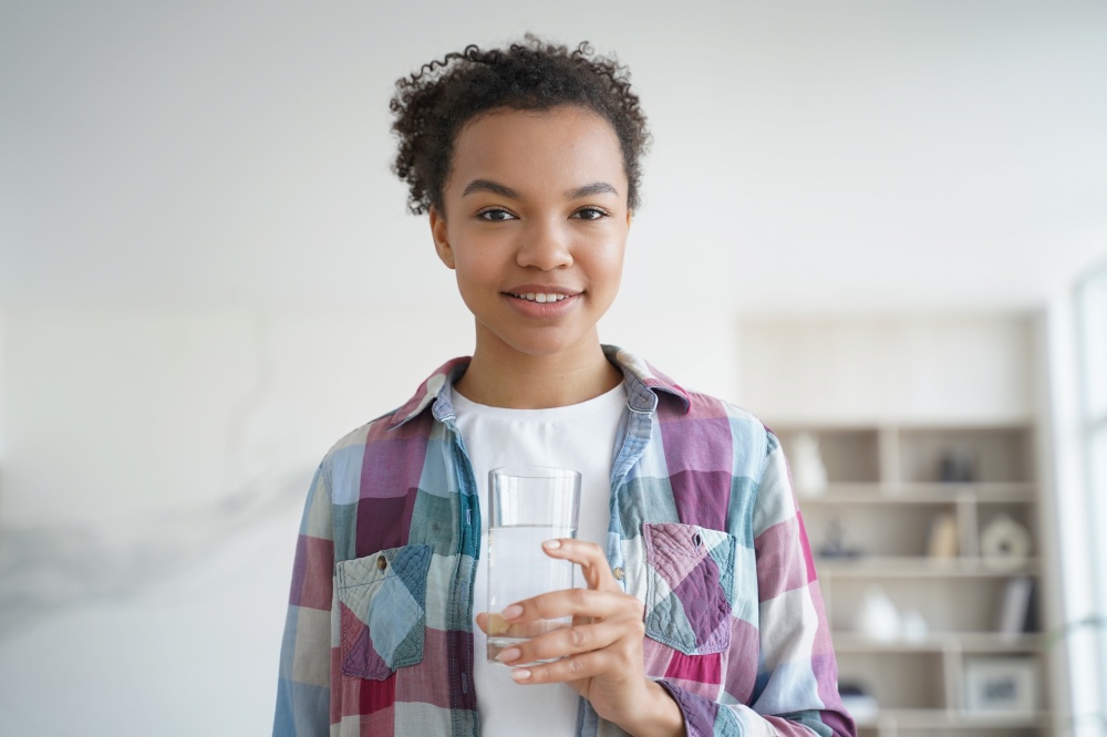 Smiling african american teen girl holds glass with mineral clean water standing at home. Happy young woman looking at camera holding fresh pure aqua. Healthy lifestyle habit, wellness concept.. African american girl holds glass of purified water standing at home. Healthy lifestyle, wellness