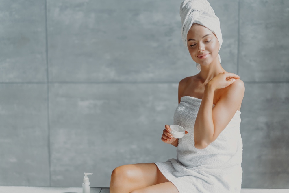 People, beauty, spa and cosmetology concept. Relaxed young European female puts body cream, touches shoulder gently, wrapped in white soft towel, closes eyes with pleasure, poses against grey wall