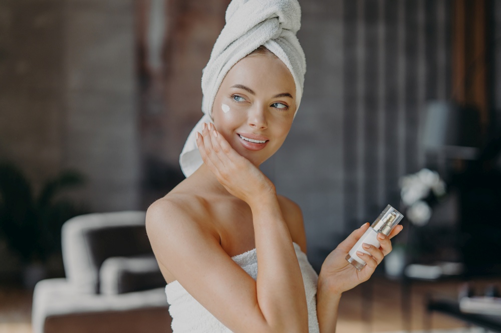 Indoor shot of young smiling woman applies moisturizer cream on face, takes care of her skin and complexion, puts lotions, has minimal makeup, wrapped in bath towel. Beauty, cosmetology concept