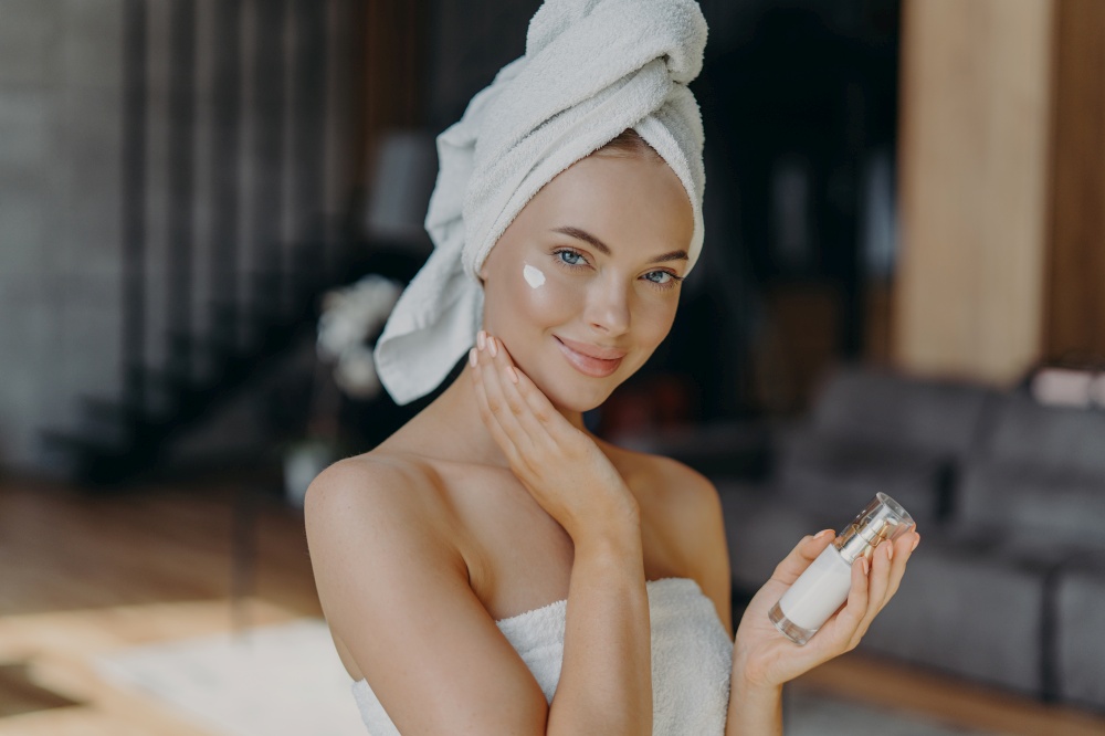 Beautiful young European woman applies face cream, hydrates skin, wears minimal makeup, wears wrapped towel on head, poses against home interior, undergoes facial treatment. Anti aginng procedure