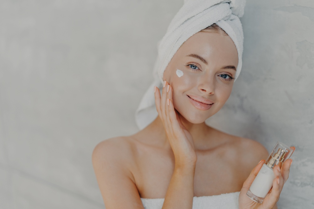 Beautiful young blue eyed woman with natural makeup applies face cream, poses bare shoulders and wrapped white bath towel on head, cares of body and complexion, poses indoor. Cosmetology concept