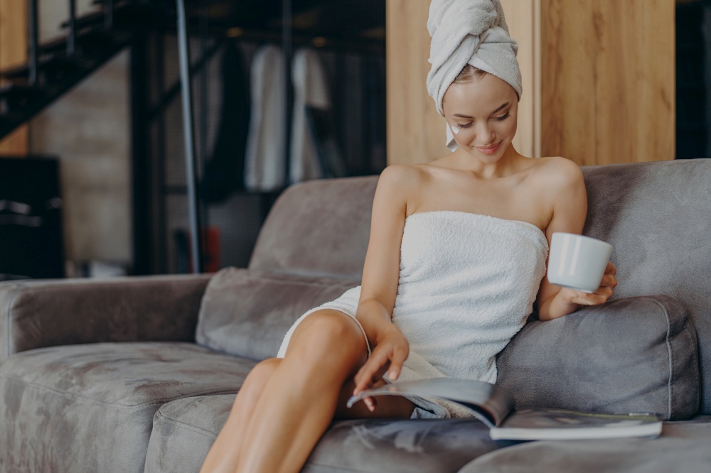 Beautiful young woman wrapped in bath towel, reads beauty magazine, drinks hot beverage, poses on comfortable couch in living room, applies face cream, has healthy smooth skin after taking shower