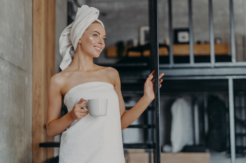 Pretty thoughtful woman concentrated somewhere, smiles broadly, has white perfect teeth, applies face cream, stands wrapped in towel, drinks hot tea, stands on stairs indoor, relaxes after taking bath