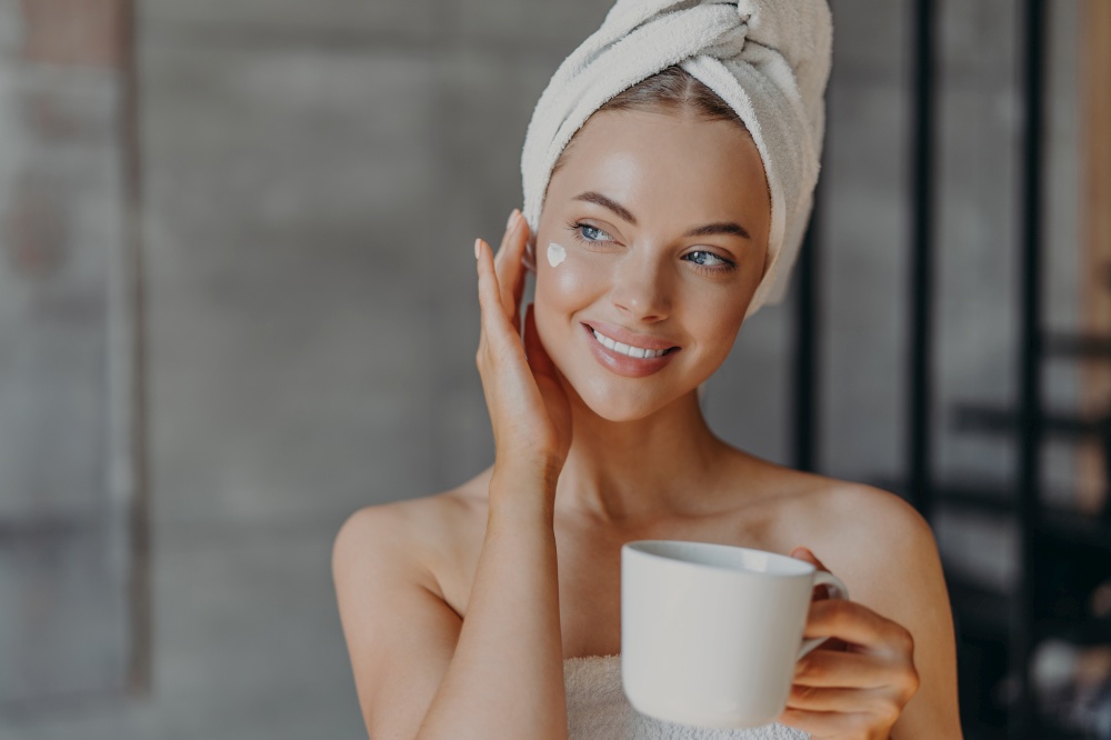 Pretty charming young woman touches face gently, applies face cream, wears minimal makeup, smiles gently, wears wrapped white bath towel on head, drinks aromatic coffee, spends free time at home
