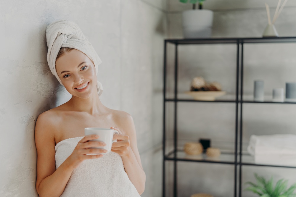 Pretty smiling woman with healthy skin wrapped in white towel, stands glad indoor, drinks coffee, has positive face expression, poses indoor. People, spa, pampering, skin care and wellness concept
