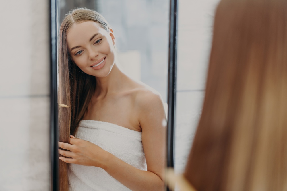 Photo of lovely woman brushes long healthy hair with comb, looks at herself in mirror, takes care of her beauty, wrapped in bath towel, poses in bathroom. Hair care and womens beauty concept