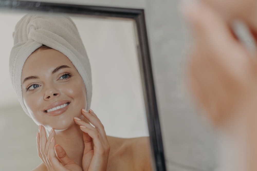 Healthy adorable young woman looks at her reflection in mirror, touches healthy glowing smooth skin, wears minimal makeup, bath towel wrapped on head after taking shower. Beauty routine concept