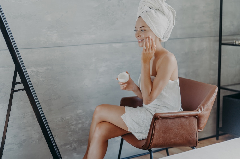Skin care concept. Indoor shot of pretty smiling woman sits in comfortable armchair opposite mirror, applies cosmetic cream on face, wears wrapped towel on head, wants to have well groomed complexion