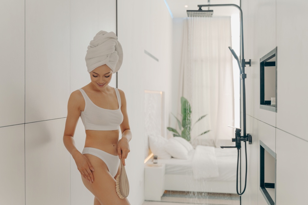Attractive young lady in white underwear doing scrubbing massage with dry brush before morning shower in light colored bathroom on background of stylish bedroom. Women beauty and hygiene concept. Beautiful young woman doing scrubbing massage in bathroom