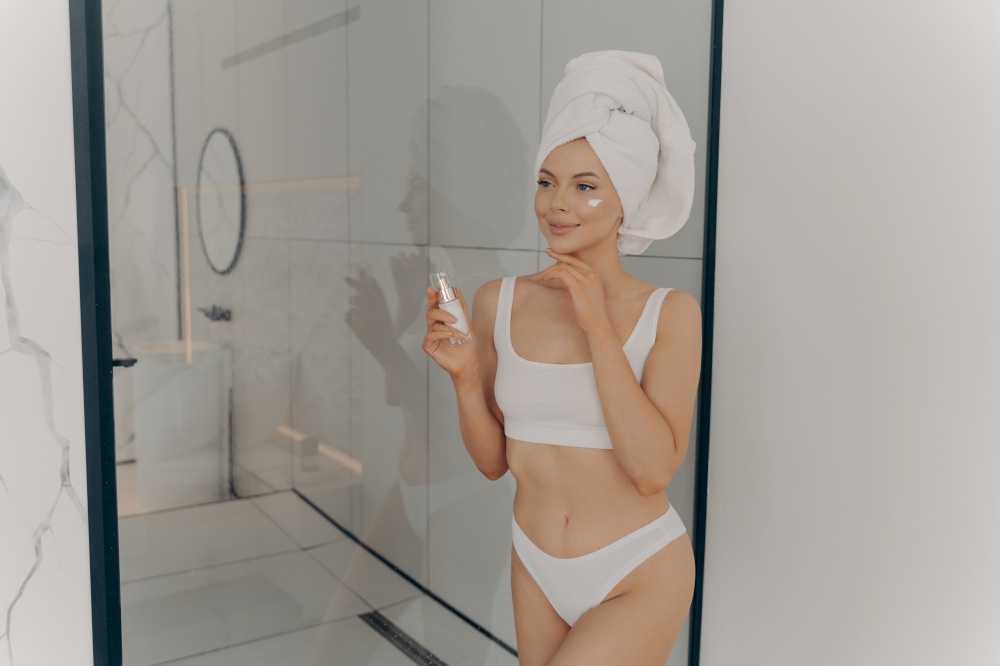 Attractive young adult woman applying facial cream, beautiful healthy lady with head wrapped in towel putting moisturizing lifting nourishing day cream on clean soft skin in bathroom at home. Attractive young adult woman applying facial cream after morning shower