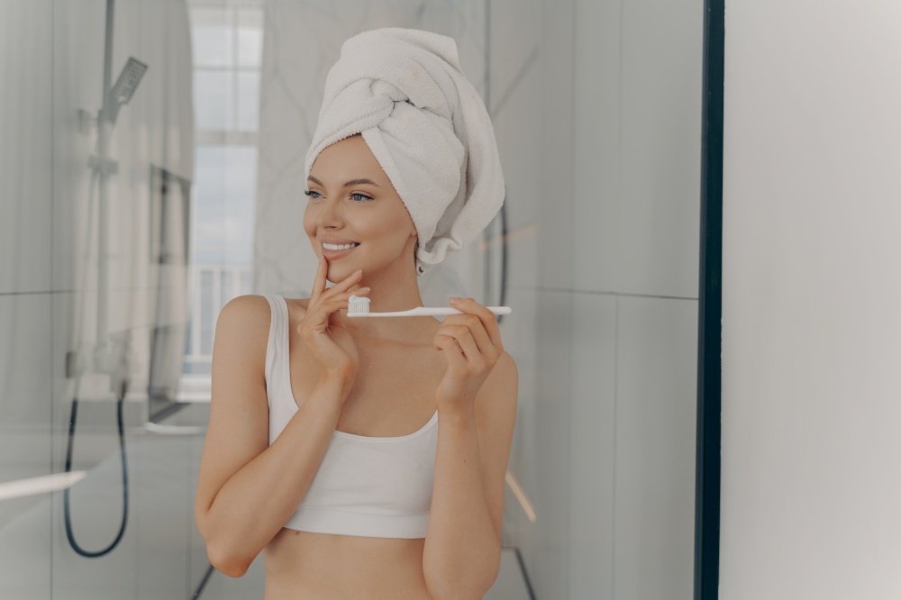 Oral hygiene concept. Healthy beautiful woman with white bath towel on head brushing teeth during morning routine, pretty young female with healthy toothy smile standing in bathroom after shower. Oral hygiene concept. Healthy beautiful woman with white bath towel on head brushing teeth during morning routine