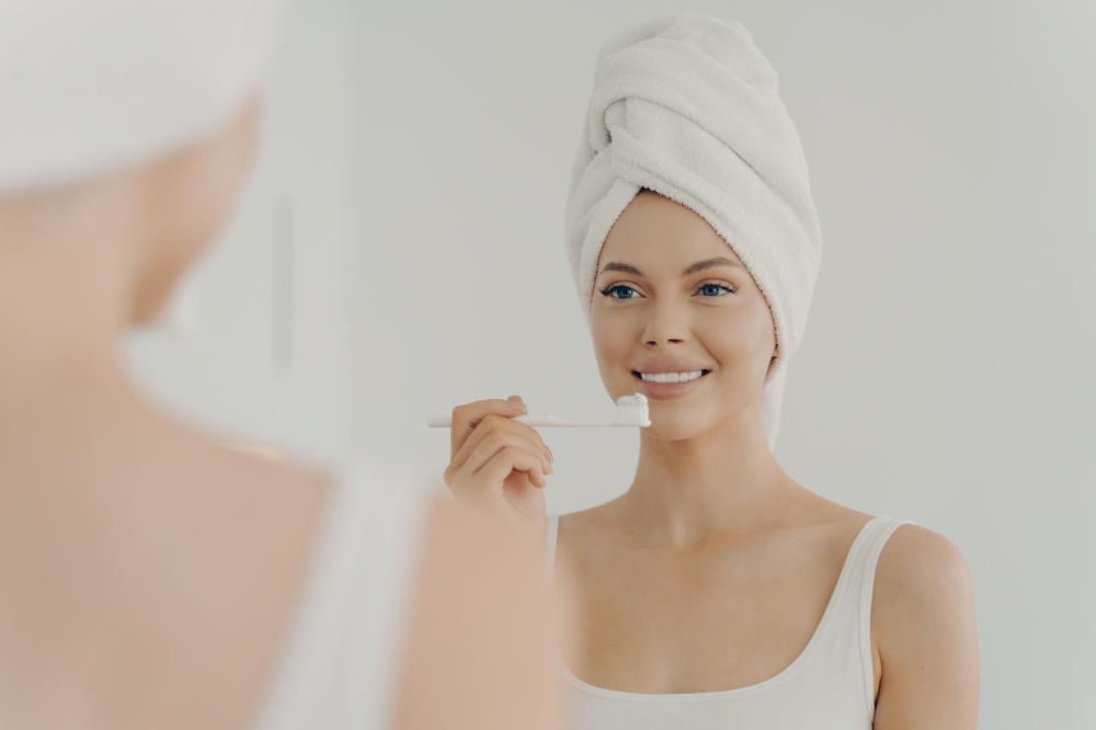 Portrait of young healthy beautiful woman with towel on head after shower having toothy smile while gently brushing her teeth, looking in mirror with pleasant facial expression. Dental health concept. Healthy beautiful woman with towel on head after shower having toothy smile while gently brushing her teeth