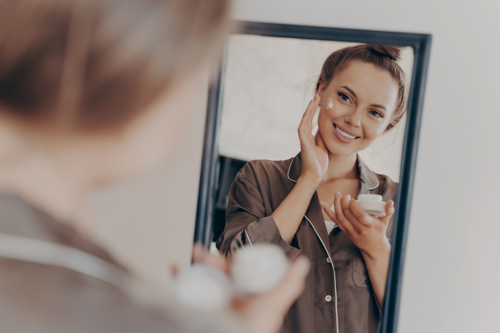 Reflection in mirror of beautiful woman in silk pajama smiling while applying face cream for deeper repair, protecting facial skin. Skincare, beauty and effective anti wrinkle treatment concept. Reflection in mirror of beautiful woman in silk pajama smiling while applying face cream for deeper repair