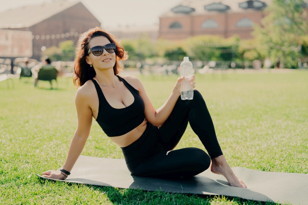 Positive active woman has perfect body shape as result of regular fitness training poses on karemat with bottle of water has rest after workout spends free time outdoors. Practicing yoga in morning