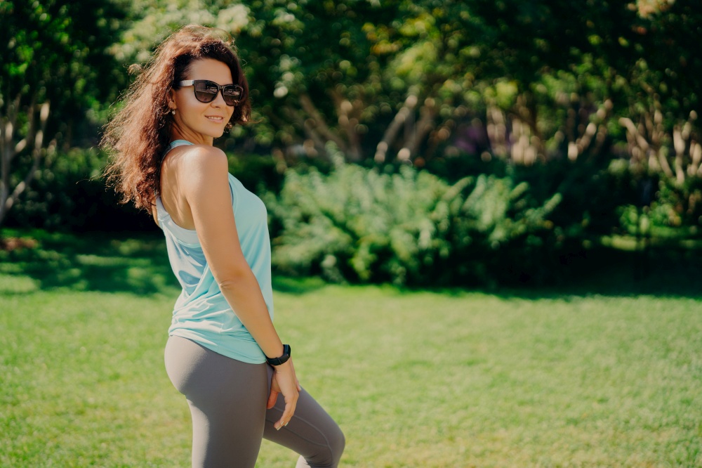 Pleased brunette European woman in active wear poses sideways wears sunglases smartwatch has fitness outdoors rests after jogging smiles pleasantly. Health body care sport and exercising concept