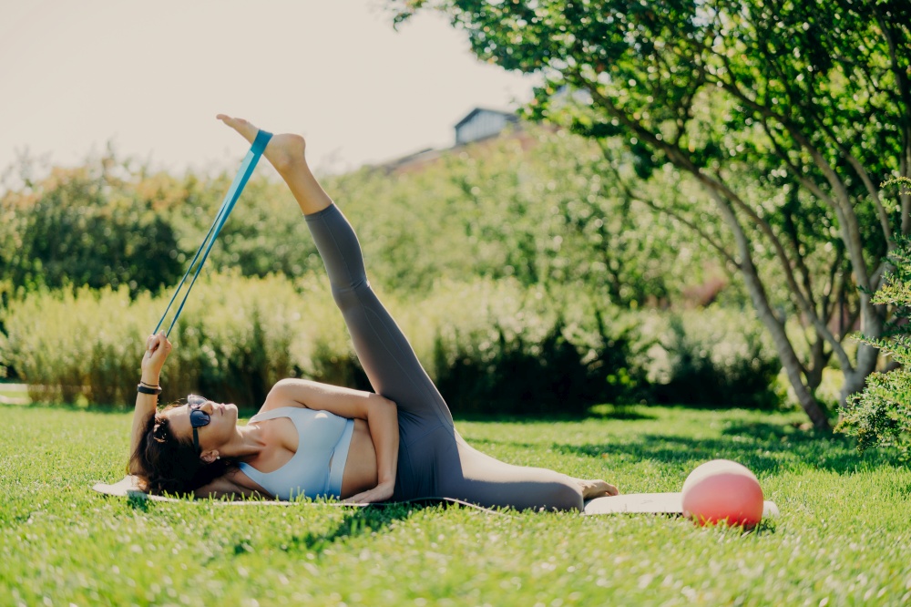Horizontal shot of flexible woman raises leg does exercises with fitness gum lying on karemat wears leggings and cropped top has perfect figure poses outside on green grass. Sportswoman uses expander
