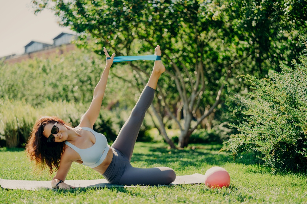 Strong fit woman uses fitness gum to stretch legs and arms stands on side plank at karemat dressed in sportswear poses outdoor during summer day everything is green. Exercises on leg muscles