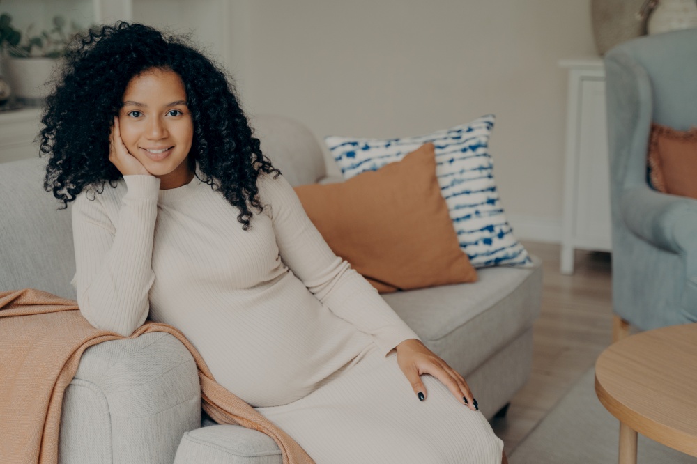 Beautiful happy african expectant mother sitting in relaxed pose while being pregnant, resting head on hand leaning on side of cozy sofa in living room, smiling and enjoying her leisure time at home. Beautiful african expectant mother sitting in relaxed pose in living room
