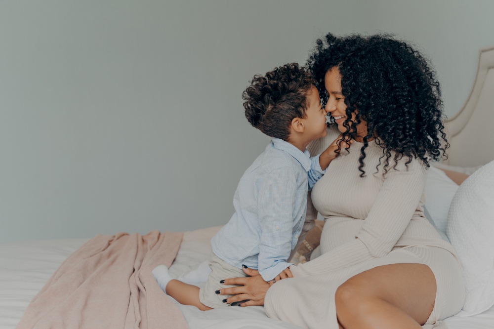 Tender moment of young family, afro american pregnant mother and son sitting on bed,touching each other with noses, white wall background in bedroom interior. Happy parenting and motherhood concept. Afro American mother and little son sitting on bed and touching each other with noses