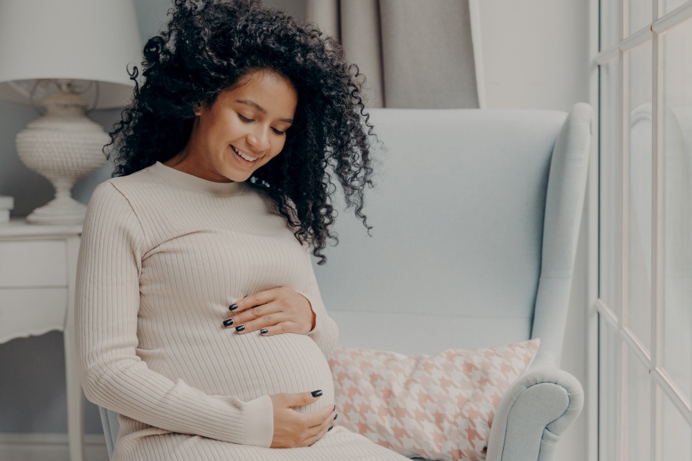 Beautiful mixed race pregnant woman in casual dress sitting in armchair by window tenderly embracing belly thinking about new baby to come. Happy maternity and pregnancy concept. Happy beautiful pregnant woman in casual dress tenderly embracing belly