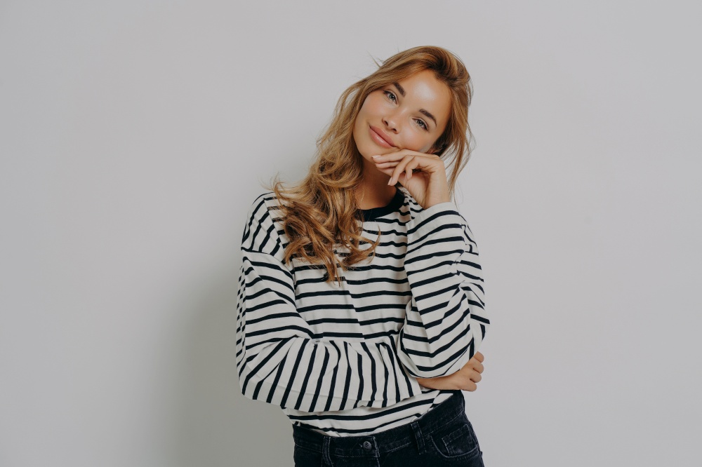 Studio shot of satisfied young European woman keeps hand on face tilts head looks gently at camera weras casual striped jumper and jeans isolated over grey background. Human face expression concept. Studio shot of satisfied young European woman keeps hand on face tilts head looks gently at camera